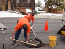 Two workers are shown digging around a sewer manhole ring and lid.