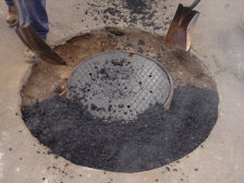 Workers are shown placing recycled asphalt around the sewer manhole ring and lid.